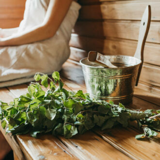 A person is sitting on a wooden bench in a sauna, with a leafy bouquet of birch twigs in the foreground.
