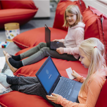 Two young girls sitting on a red couch with laptops in their laps.