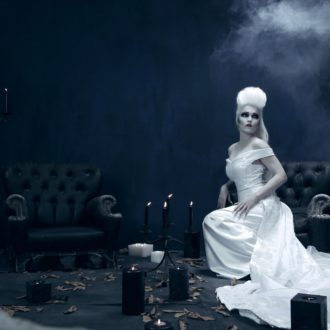 Tarja Turunen in a white dress and wig sitting on a black armchair amidst candles.