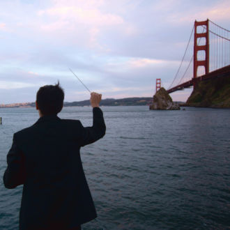 A man, his back to the camera, waves a conductor's baton on the shore by the Golden Gate bridge.