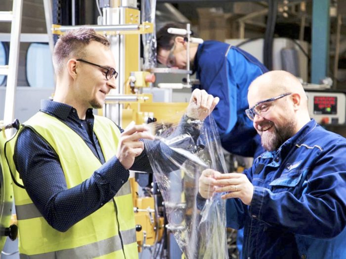 En un taller, dos hombres vestidos con ropa de trabajo y gafas sostienen una lámina de un material que parece plástico tranparente para envoltura.
