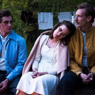 Two men and one woman sitting outside in a row. The men make eye-contact while the woman is leaning on the other man.