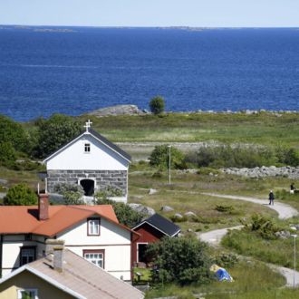 Several wooden houses sit among the hills of an island, with the ocean and the sky visible in the background.