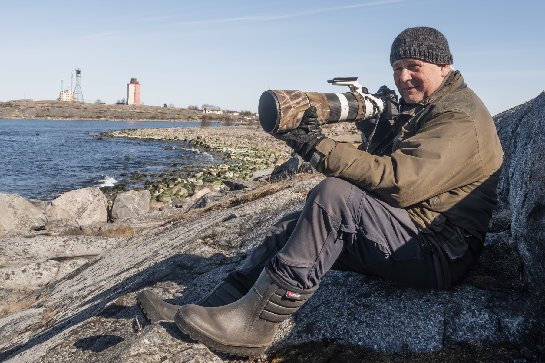 A man sits on a rocky ocean shoreline and adjusts a camera with a long lens.