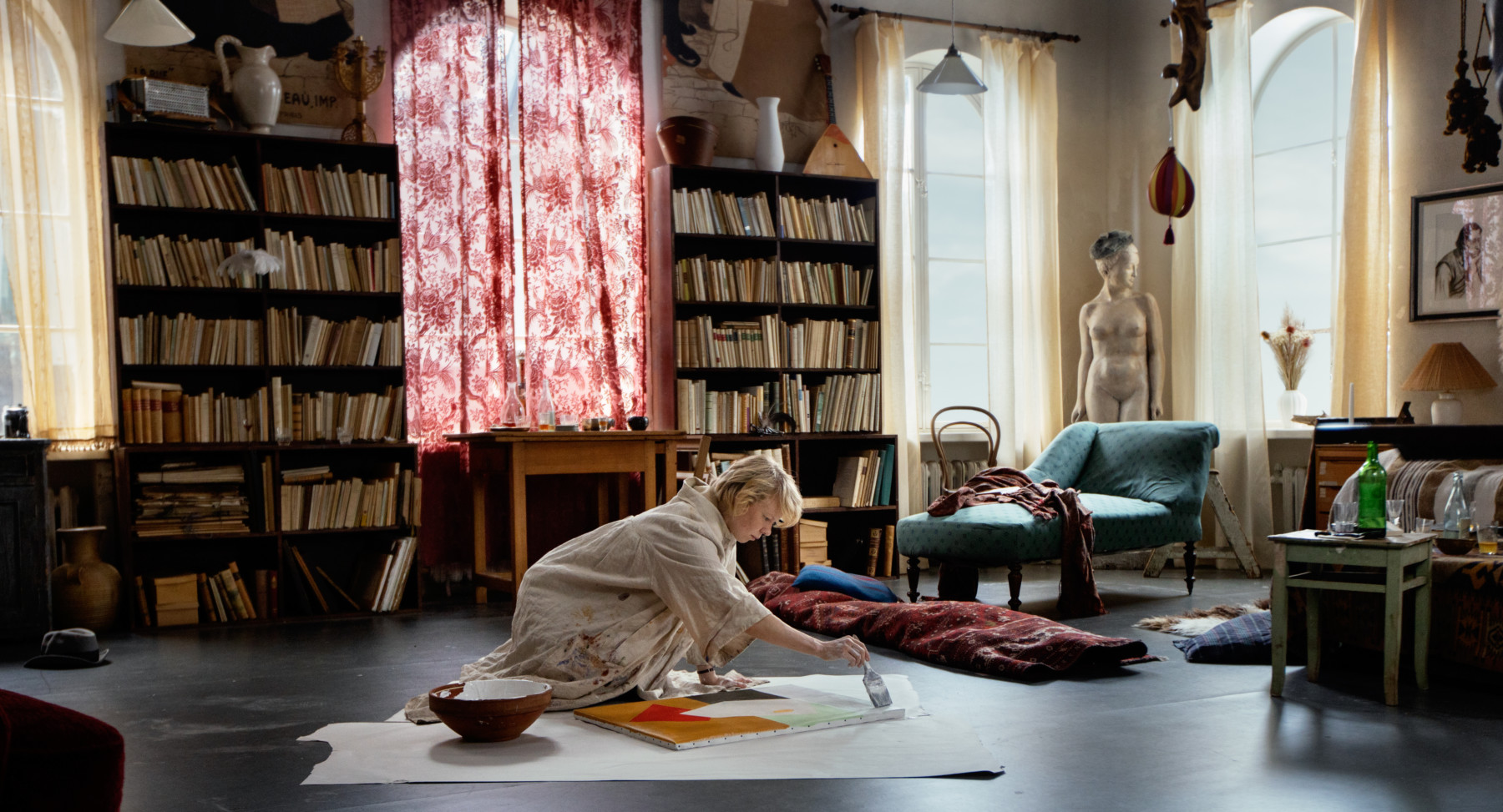 A woman paints on a canvas on the floor of a large room with bookshelves in the background.