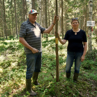 Um homem e uma mulher estão parados na floresta, cada um com uma das mãos em um alto mastro de madeira cravado no chão.
