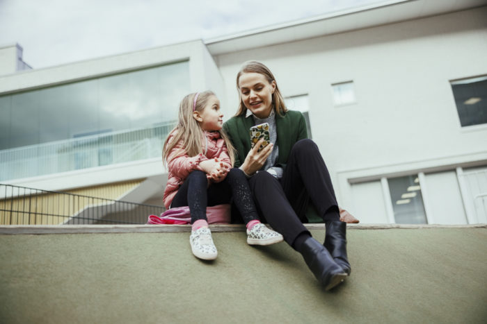 A woman and a girl sit outside in front of a school building, looking at a phone the woman is holding.