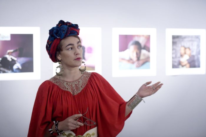 A woman gestures with her hand in front of a museum wall on which large framed photos are visible.