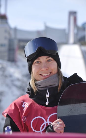 A woman dressed in sporty winter clothing holds a snowboard and smiles at the camera.