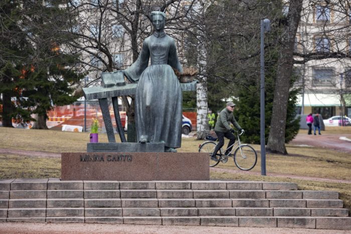 A statue of a woman holding an open book stands in a park while a man bikes past in the background.