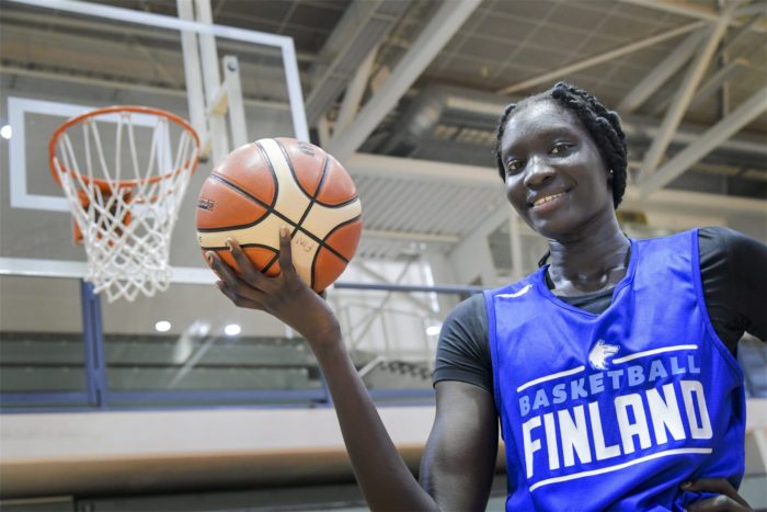 A smiling young woman in a shirt with the word “Finland” on it holds a basketball in her upturned palm.