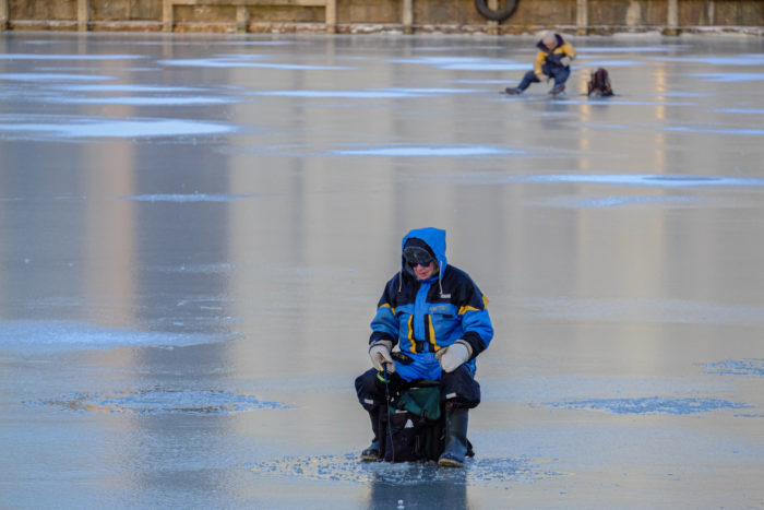 Two people in winter clothes sit on stools on a wide expanse of sea ice.