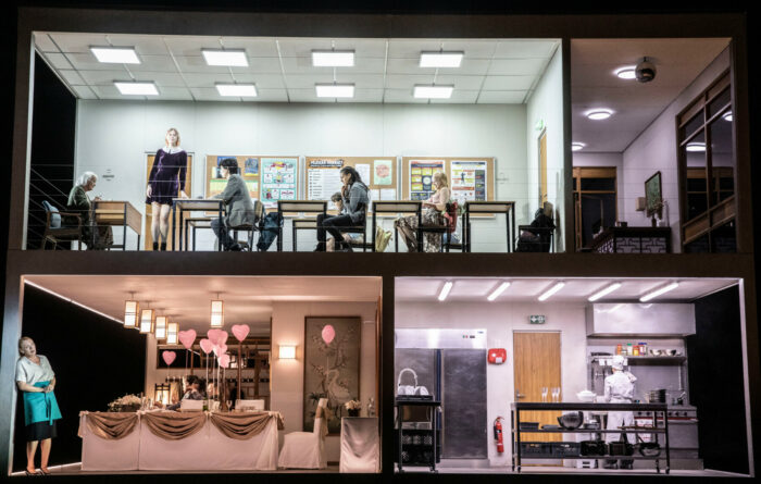 On a multilevel stage set, singers stand and sit in spaces designed to look like the rooms of a school and a restaurant.