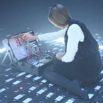 A woman in a black dress and white shirt sits in front of an audio mixing board, on a floor covered with VHS cassettes.