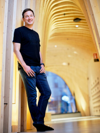A man in blue jeans and a black T-shirt leans against the wall in a hall featuring wooden archways.