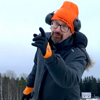 A man dressed in heavy winter clothing holds up one finger and smiles.