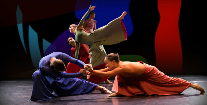 Cinq ou six danseurs et danseuses habillés de costumes de scène de couleurs claires se tiennent en équilibre dans des poses variées.