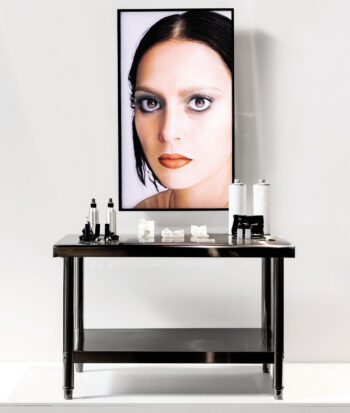 A makeup table with bottles and implements on it, and instead of a mirror, a rectangular screen showing a woman’s face.