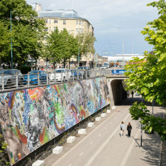 A long painting is standing along one side of a tree-lined path where two people are walking.