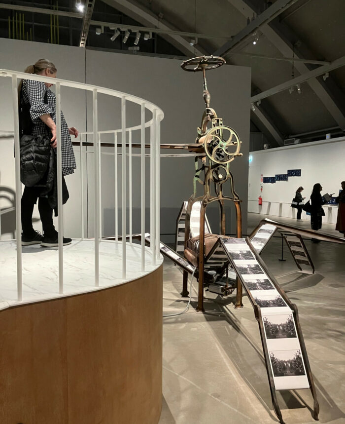 A person stands on an observation terrace looking at a large sculpture that contains parts of machines and resembles the form of a spider.