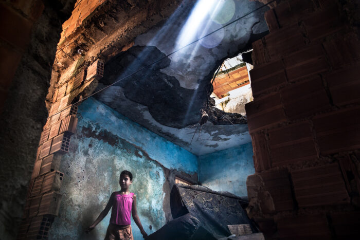 Sunlight streams in over a child of about ten years old who is standing in a half-destroyed building where there is a jumble of old furniture, a gaping hole in one wall and a hole in the ceiling.