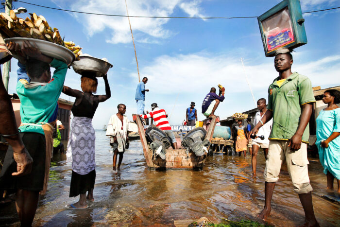People are walking with large bowls full of branches and other goods on their heads, through an area covered with several centimetres of water.