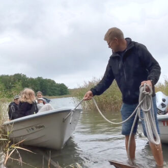 A man stands ankle-deep in water on a shoreline while pulling on a rope attached to a small boat where several children are sitting.