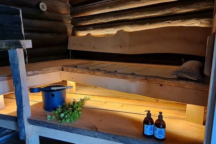 Um banco de sauna em madeira contém um balde e um buquê de ramos de bétula, numa sauna com paredes de troncos.