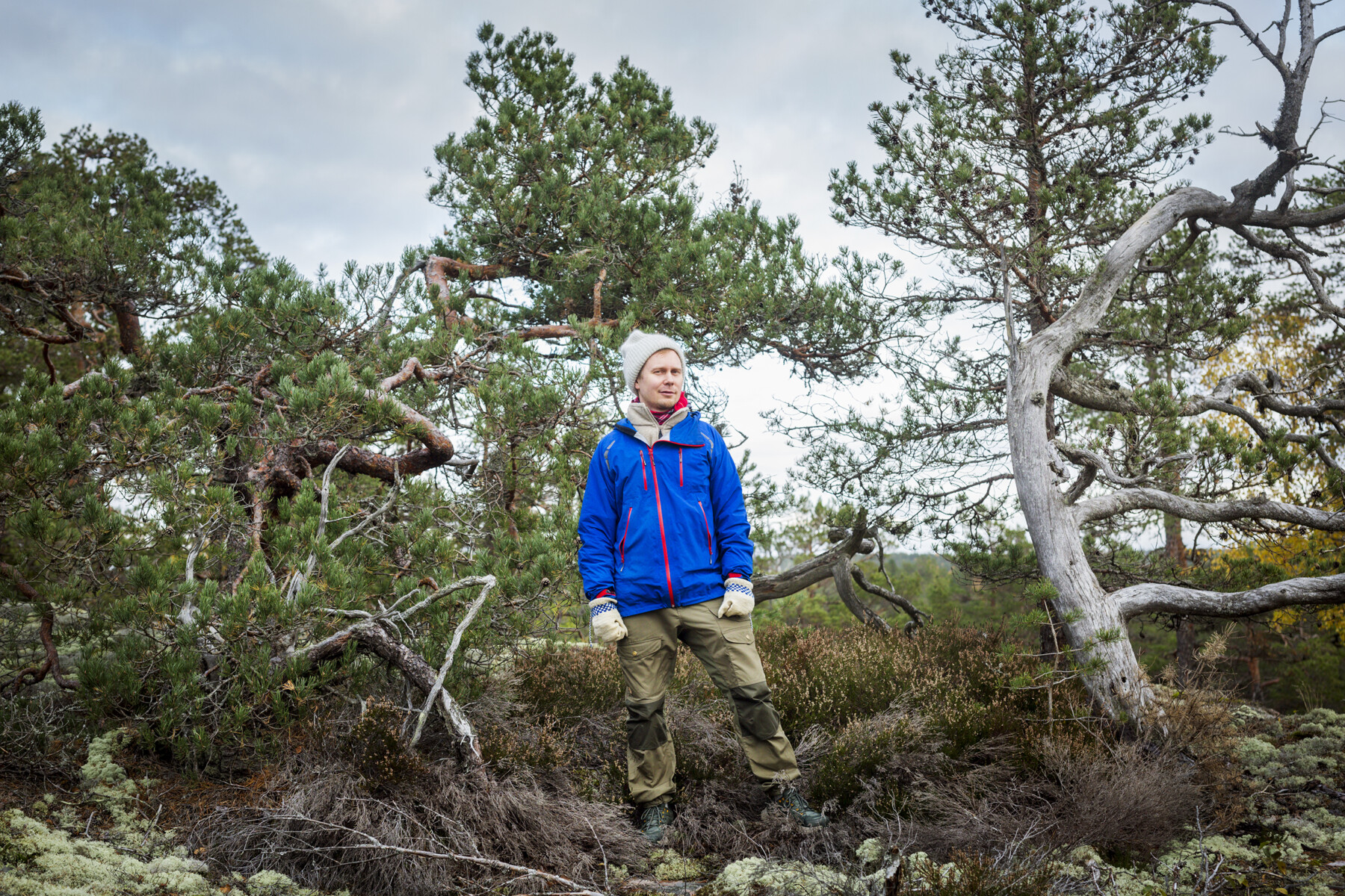 A man is standing in a forest in clothing that show that the weather is chilly.