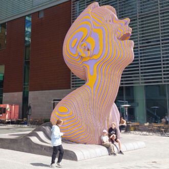Under a roof made of glass and metal, people sit at the base of a sculpture that is about four times as tall as they are, depicting a head looking upwards with an open mouth.