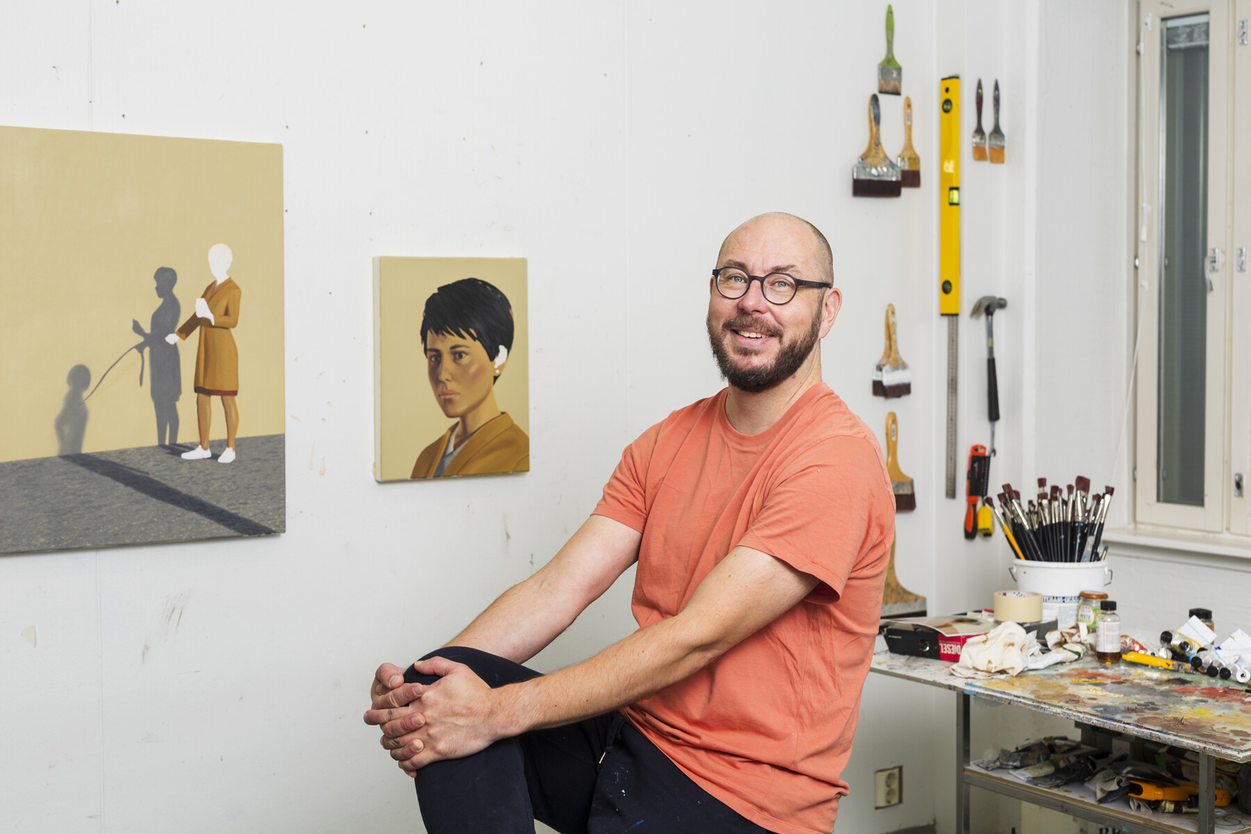A man is sitting in an art studio with brushes, tools and paintings hanging on the wall.