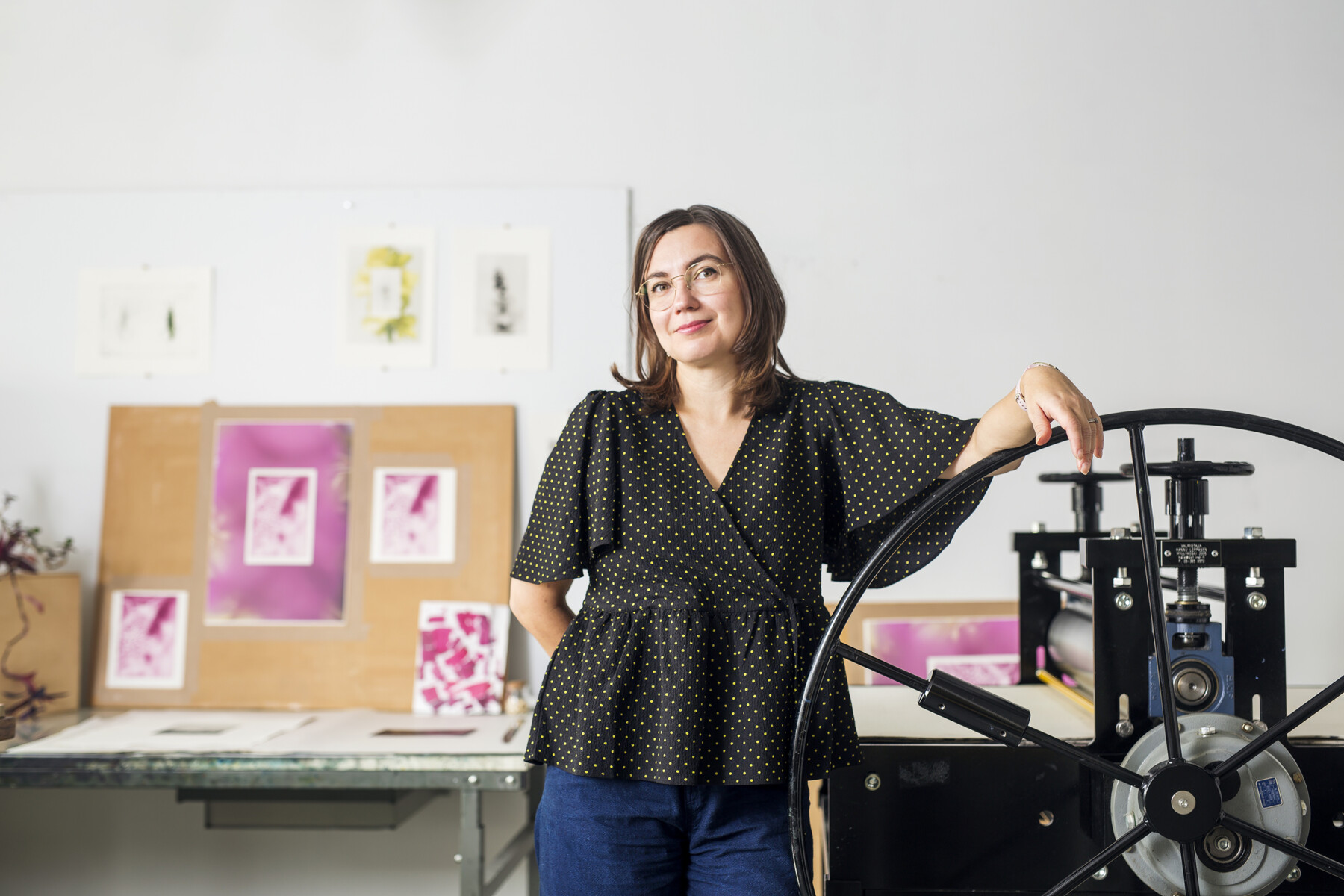 In an art studio with prints hanging on the wall, a woman rests one arm on the crank wheel of an old-fashioned printing press.