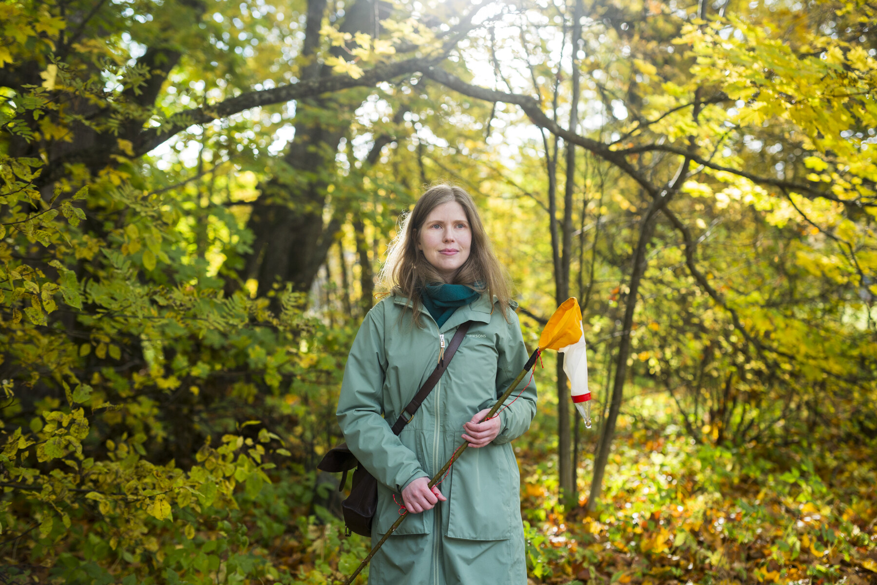 A woman is standing in a forest holding a small net on a long handle.