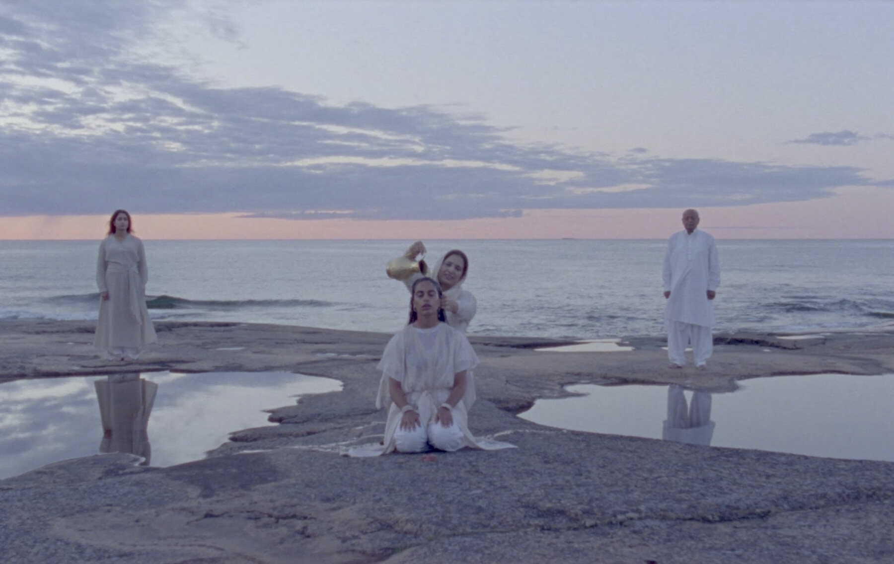 On a coast among tidepools, a woman is standing and pouring water over the hair of a kneeling woman, while two other people stand in the background.
