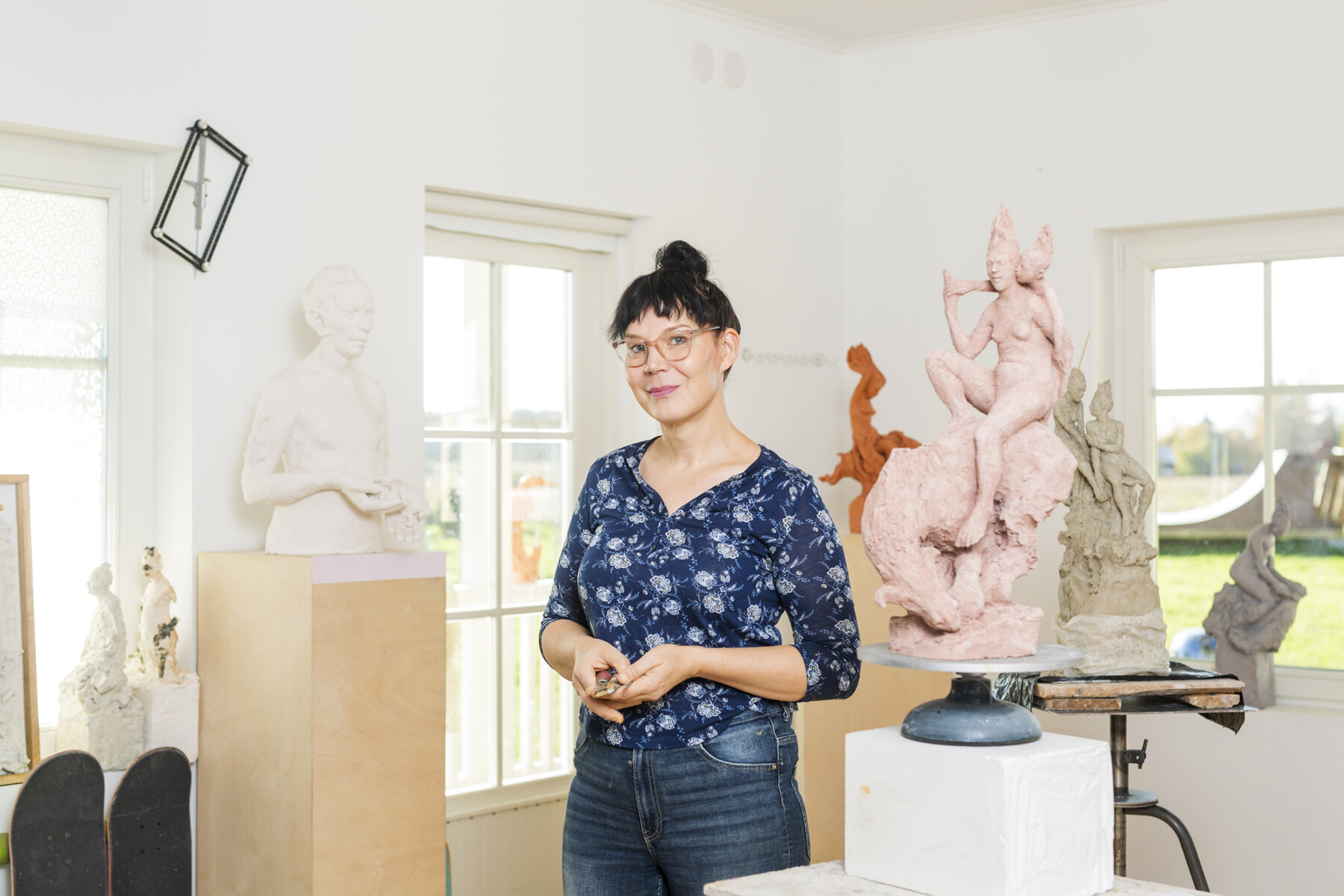 A person stands in an art studio or exhibition space with several sculptures on pedestals, a couple skateboards, and large windows overlooking a yard with a skateboard ramp.