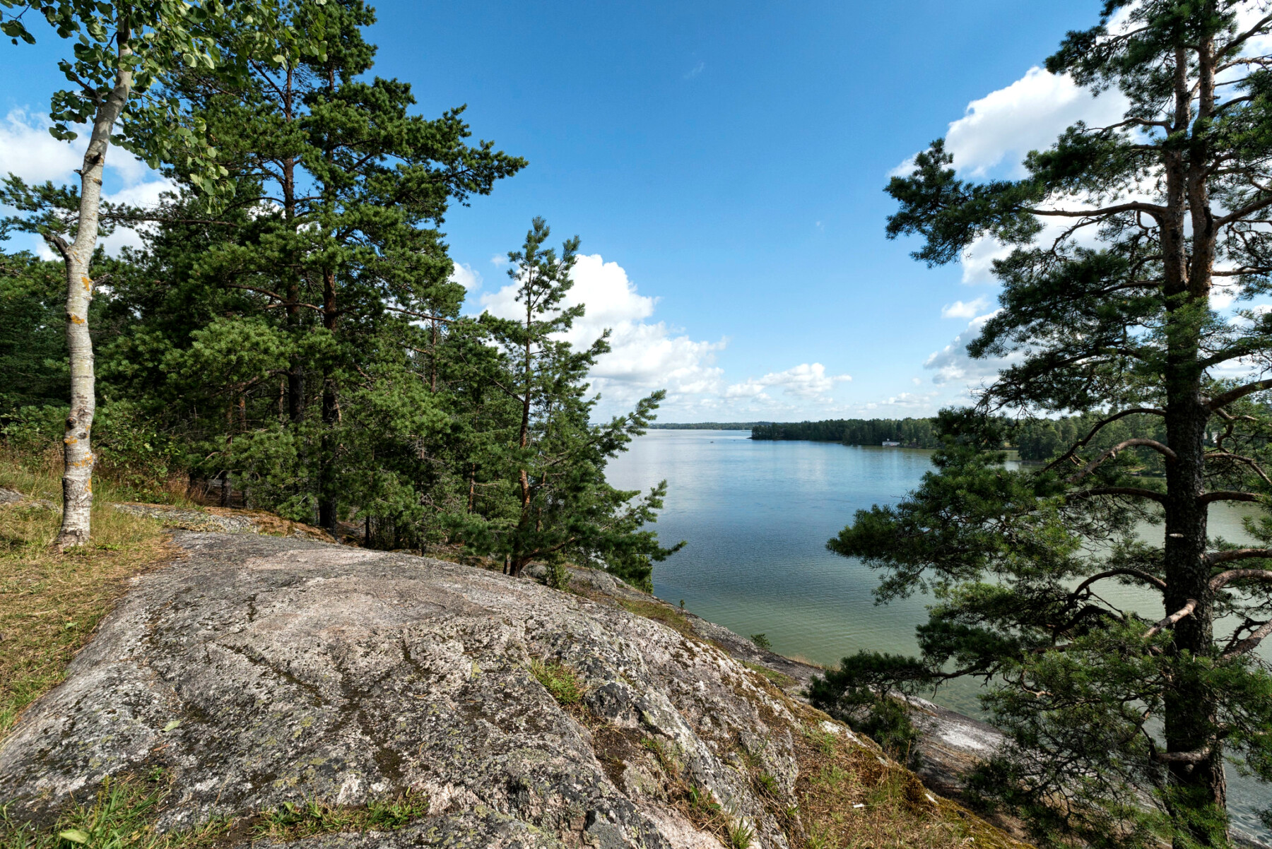 A blue lake and a blue sky are visible beyond a large boulder and several trees.