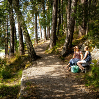 Two women in summer attire are sitting on a bench in a forest by a lakeside path.