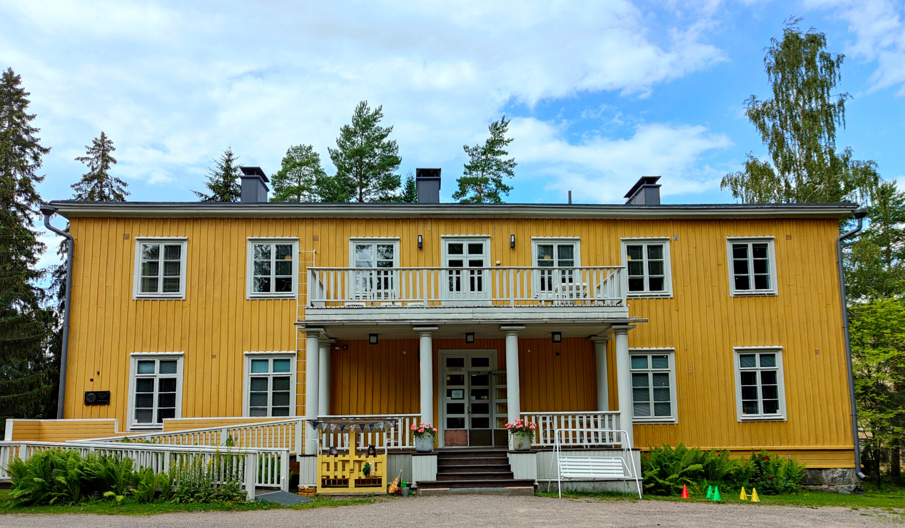 A long, two-storey yellow wooden house with a veranda is flanked by trees.