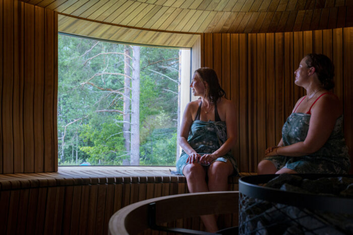 Two women wrapped in towels sit on a bench in a sauna, looking out the window at a forest.