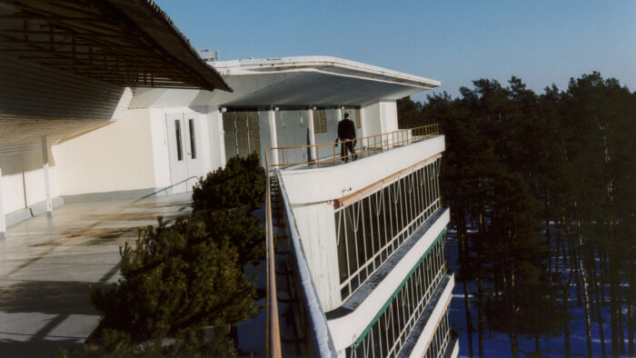 Eine Person läuft im obersten Stockwerk eines Gebäudes eine Terrasse entlang, den Hintergrund bilden Baumkronen.
